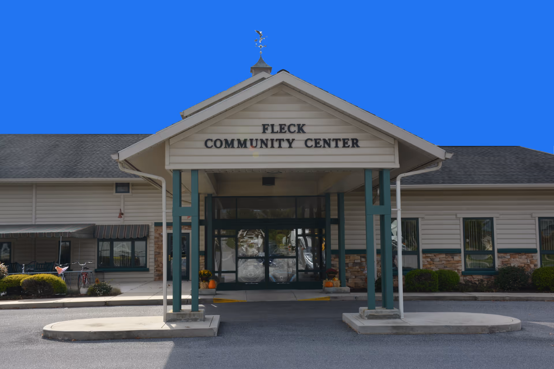 Exterior view of the Fleck Community Center building entrance with a covered drop-off area supported by green pillars. The building has beige siding with stone accents and several windows. There are some bushes and a bicycle visible on the left side near the entrance.