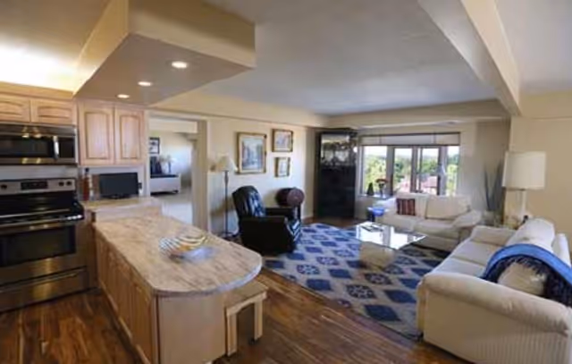 Open-plan interior showing a kitchen island and adjoining living area with sofas, a recliner, rug, and large window.