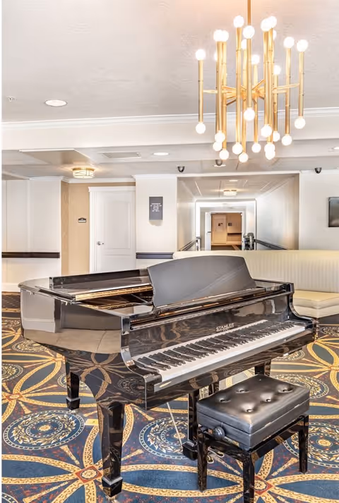 A black grand piano with a matching padded bench sits on a patterned carpet in a well-lit room. Behind the piano, there is a cream-colored curved sofa and a hallway leading to other rooms. A modern chandelier with multiple light bulbs hangs from the ceiling.