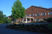 Exterior view of a two-story brick building with a covered entrance, surrounded by landscaped bushes and trees under a clear blue sky.