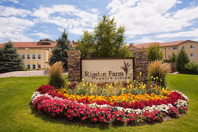 A landscaped garden area with colorful flowers including red, white, yellow, and orange blooms surrounding a stone and wood sign that reads 'Rigden Farm Senior Living'. Behind the sign are trees and a large building with a red roof under a partly cloudy sky.