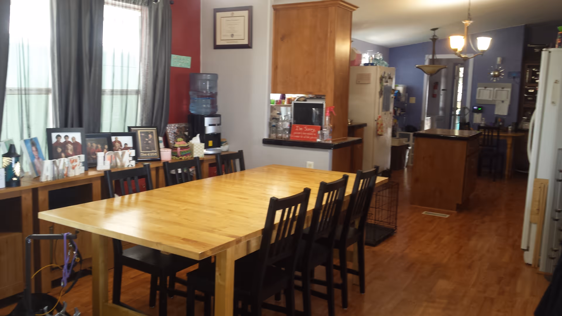 Interior view of a dining area with a large wooden table surrounded by black chairs. In the background, there is a kitchen area with wooden cabinets, a refrigerator, and a water dispenser. Family photos and decorative items are displayed on a sideboard against a wall with windows covered by curtains.