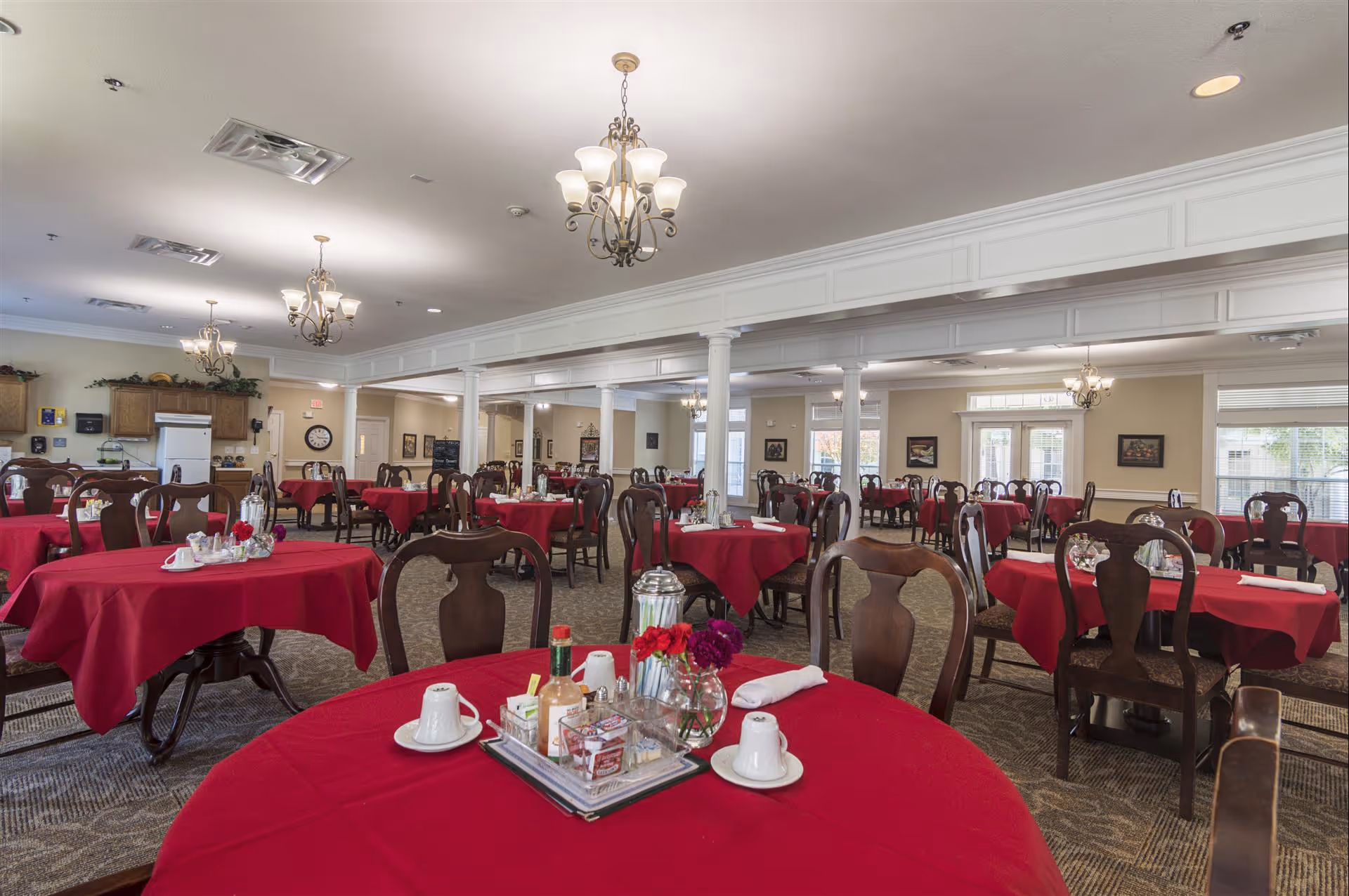 Spacious dining room with multiple round tables covered in red tablecloths, place settings, and wooden chairs under chandeliers.