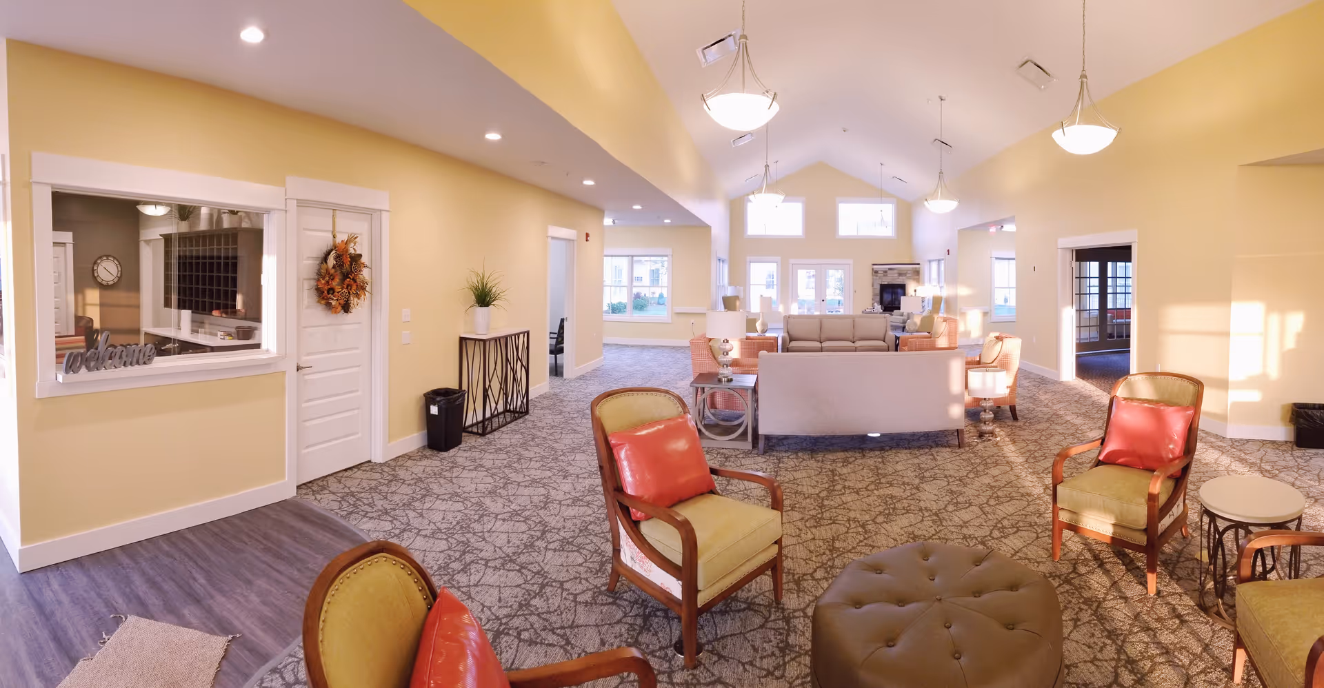 A spacious and well-lit living room area in an assisted living community with yellow walls, carpeted floors, and multiple seating arrangements including armchairs with red cushions, sofas, and small tables. There are large windows letting in natural light, pendant lights hanging from the ceiling, and a decorative wreath on a door near a reception window with a 'welcome' sign.