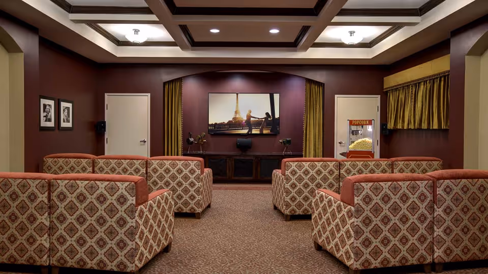 A cozy senior living facility theater room with patterned armchairs arranged in rows facing a large flat-screen TV mounted on a burgundy wall. The room features a popcorn machine on the right side, framed black and white photos on the left wall, and gold curtains flanking the TV. The ceiling has recessed lighting and decorative beams.