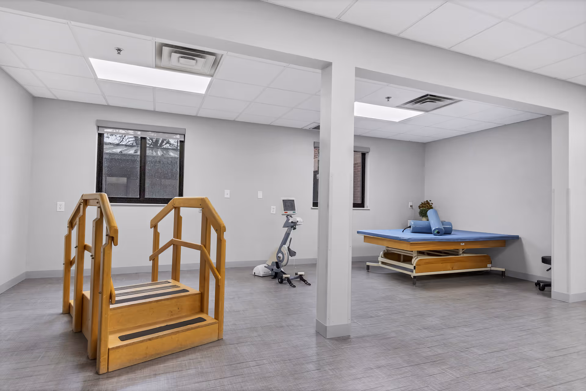 A rehabilitation or physical therapy room with a wooden set of stairs for practice, an exercise bike, and a padded therapy table with blue cushions. The room has gray walls, two windows, and a tiled floor.