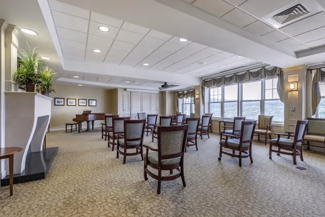 A spacious room with multiple wooden chairs arranged in rows facing a wall with a piano and framed pictures. The room has large windows with drapes allowing natural light to enter, a carpeted floor with a patterned design, and a ceiling with recessed lighting and ventilation panels.