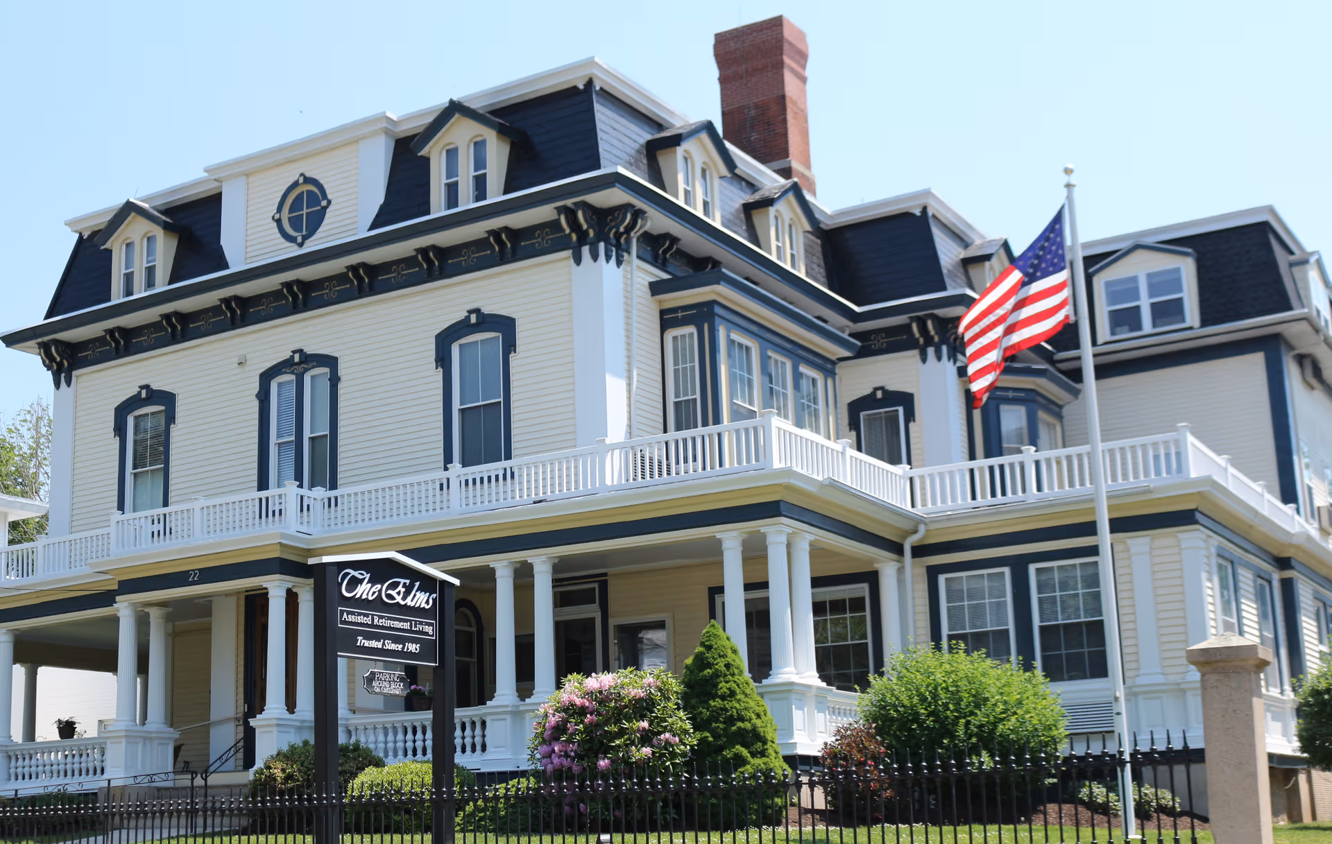 Large, elegant multi-story building with white and dark blue trim, featuring a wrap-around porch with white columns, an American flag on a flagpole in front, and a sign that reads 'The Elms Assisted Retirement Living Trusted Since 1985'. The building is surrounded by a black metal fence and landscaped greenery under a clear blue sky.