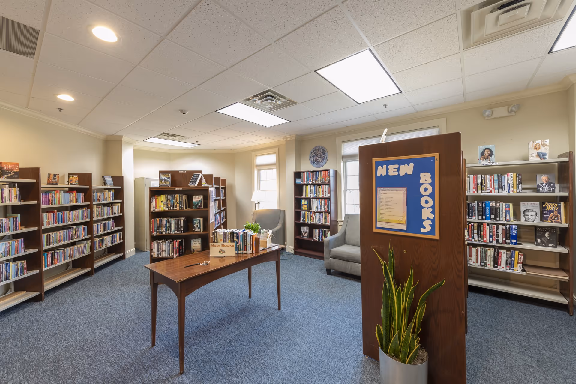Interior view of a library room in Coburg Village with multiple wooden bookshelves filled with books, a table in the center displaying new books, two gray armchairs near the windows, and a wooden divider with a blue bulletin board labeled 'NEW BOOKS'. A potted plant is placed in front of the divider.