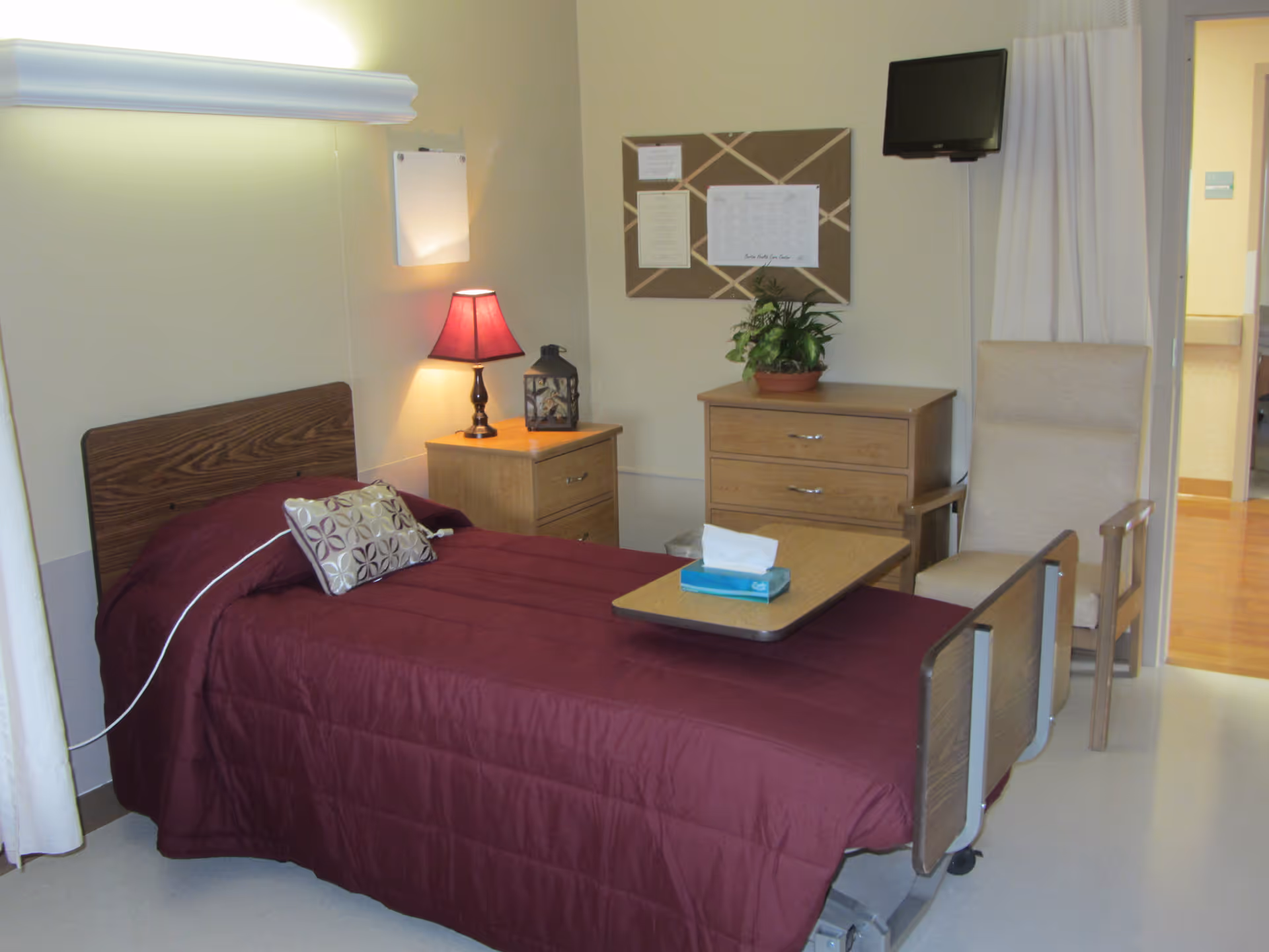 A single bed with a maroon bedspread and a decorative pillow in a healthcare facility room. Next to the bed is a wooden nightstand with a red lamp and a decorative lantern. A wooden dresser with a potted plant and a bulletin board with papers is on the wall. A beige armchair is positioned near the dresser, and a small TV is mounted on the wall above it. The room has light-colored walls and flooring, with a curtain partially visible on the left side.