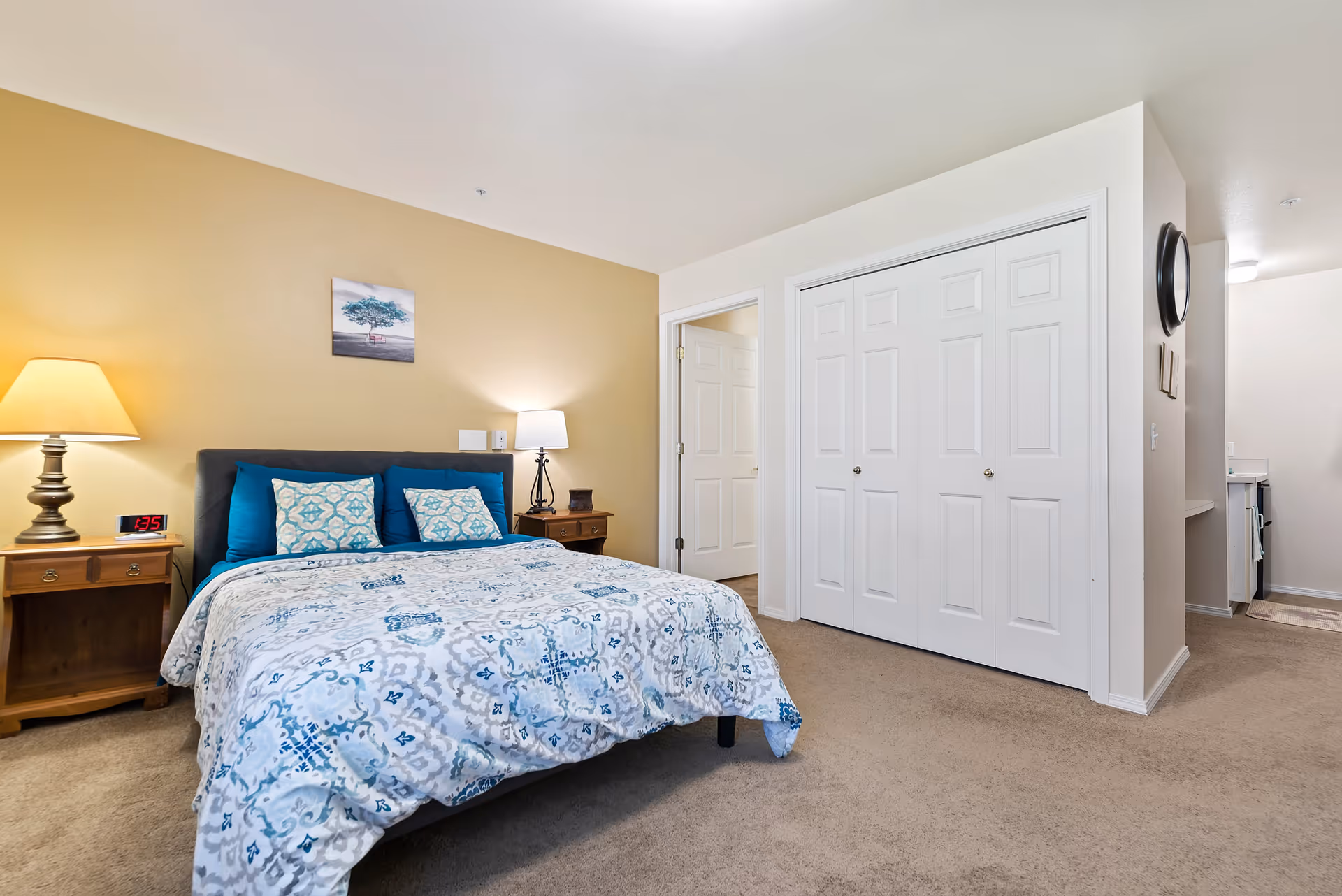 A cozy bedroom with a bed covered in a blue and white patterned comforter and matching pillows. There are two wooden nightstands on either side of the bed, each with a lamp. A digital clock on the left nightstand shows the time 1:35. The walls are painted beige and white, with a small framed picture of a tree above the bed. There is a large white closet with double doors and an open doorway leading to another room. The floor is carpeted in a light brown color.
