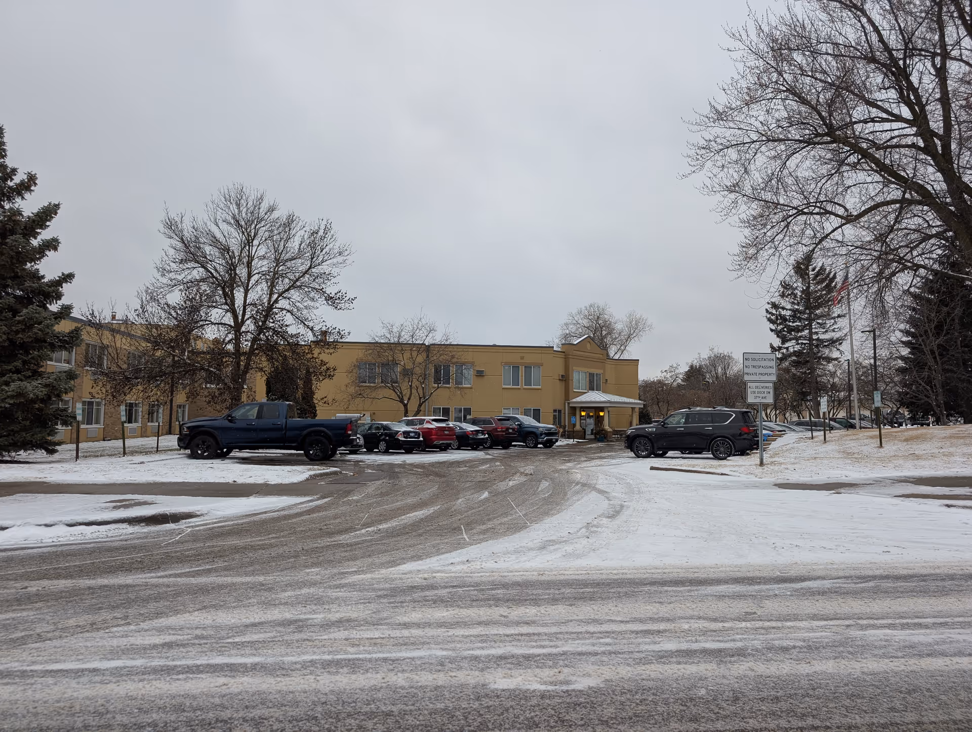 Two-story beige senior living building with parked cars, leafless trees, and a snowy driveway under an overcast sky.
