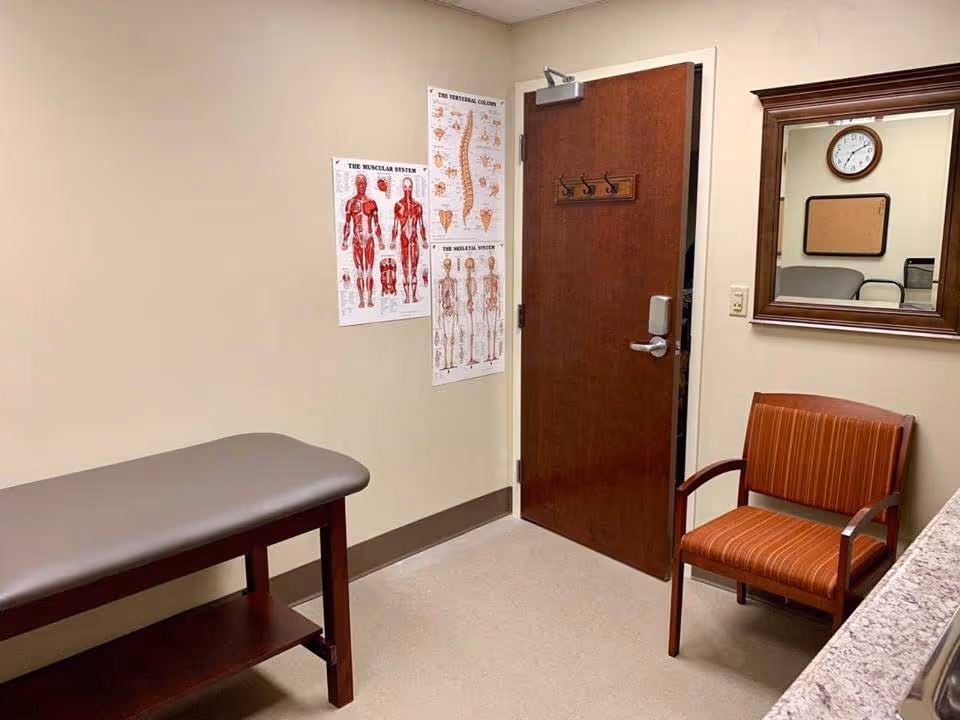 A small medical examination room with a padded examination table on the left, anatomical charts of the muscular system, vertebral column, and skeletal system on the wall, a wooden door with hooks, a striped orange chair, and a mirror reflecting a clock and bulletin board.