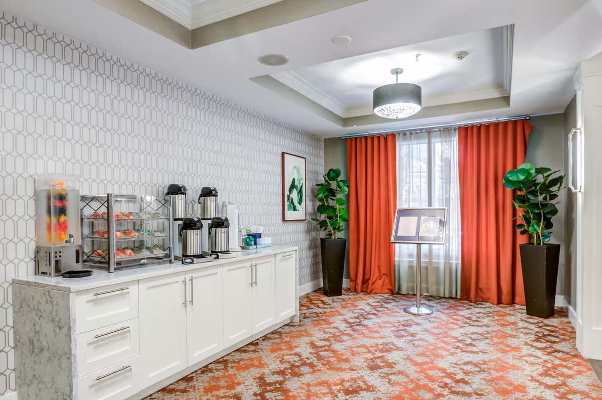 A well-lit interior space with a white cabinet topped with a marble surface holding coffee dispensers, a juice dispenser, and a display case with snacks. The wall behind has a geometric patterned wallpaper. There are two tall potted plants flanking a window with red curtains and sheer white drapes. A ceiling light fixture is centered above, and a stand with a framed document is placed in front of the window.