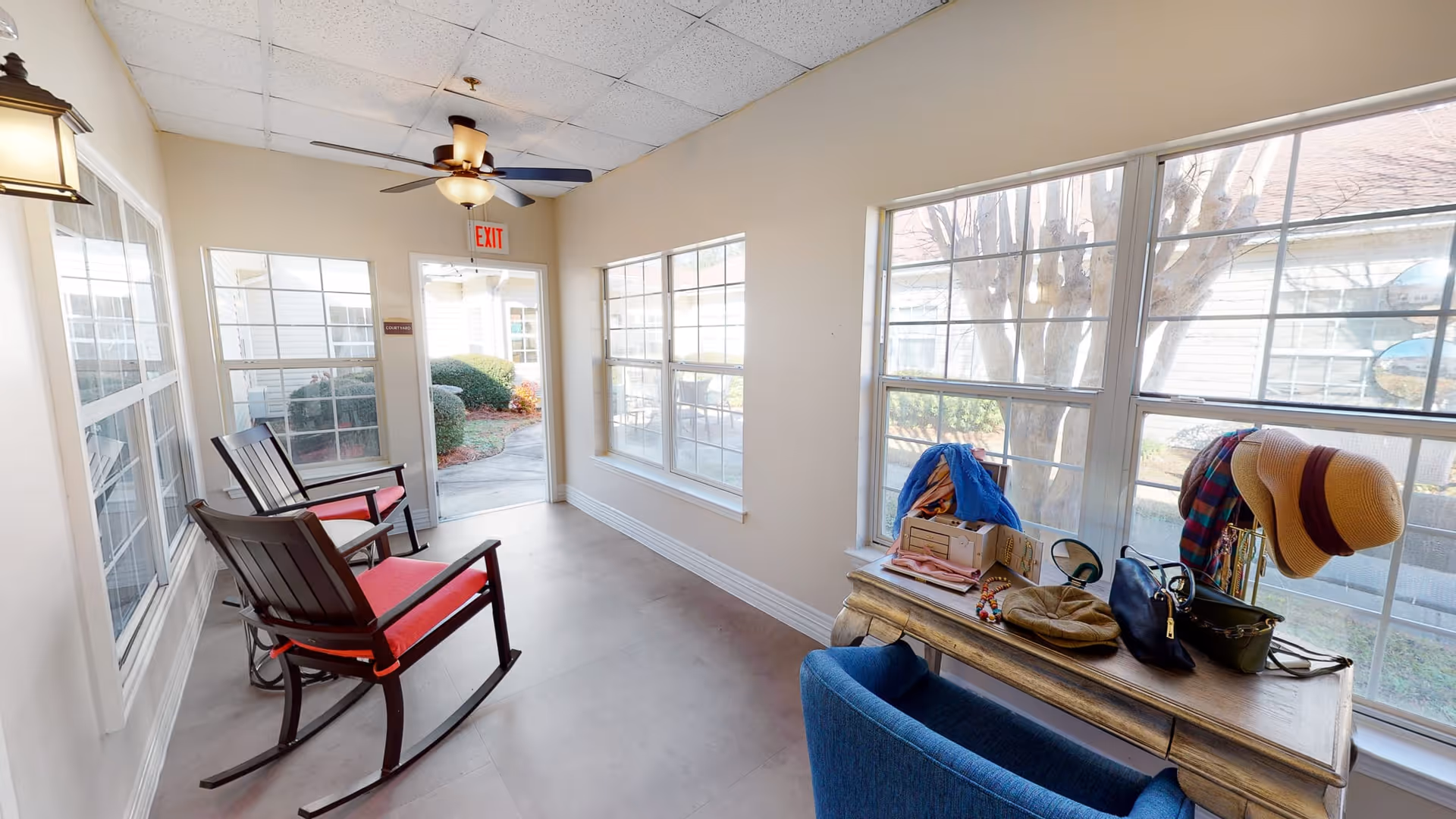 A bright sunroom with large windows on two sides, featuring two wooden rocking chairs with red cushions on the left and a wooden table on the right holding various personal items including hats, bags, and a blue jacket. A ceiling fan with a light hangs from the ceiling, and an exit door leads outside to a garden area.