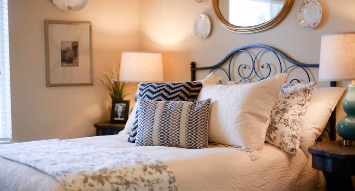 A cozy bedroom with a neatly made bed featuring multiple pillows in various patterns. The bed has a decorative metal headboard. On either side of the bed are wooden nightstands with lamps and decorative items, including a framed photo and a plant. The wall behind the bed has a round mirror and decorative plates.