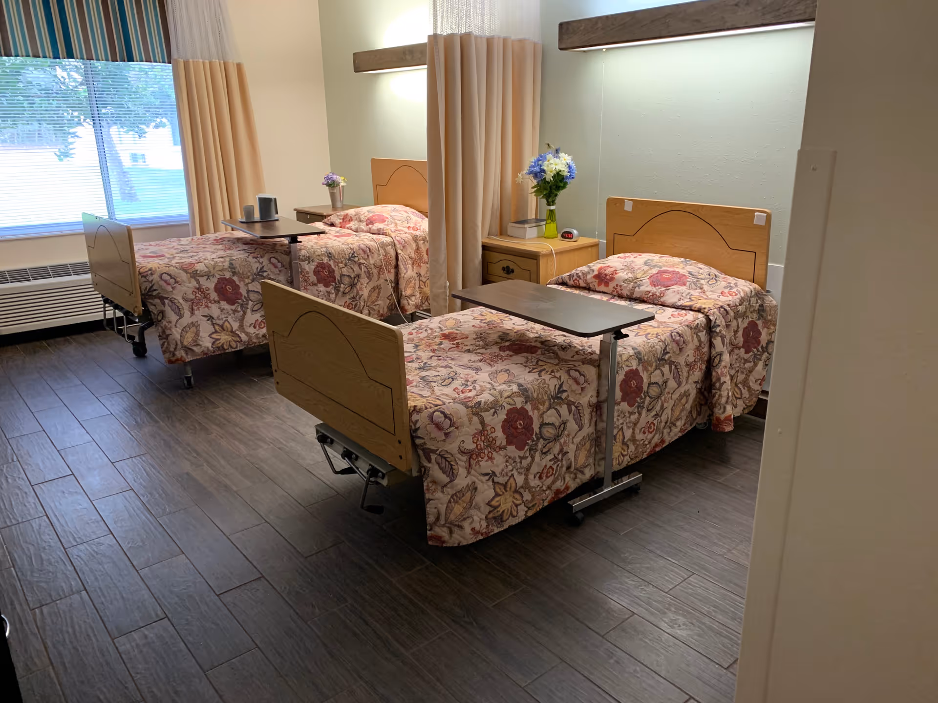 A nursing home bedroom with two hospital-style beds covered in floral bedspreads. Each bed has an overbed table and a nightstand with a vase of flowers. There is a large window with striped and beige curtains, and the floor is dark wood.