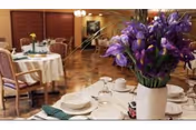A dining room set up with round tables covered with white tablecloths, each table set with plates, cups, and silverware. There are wooden chairs with cushions around the tables. In the foreground, there is a white vase filled with purple flowers. The room has wooden paneling on the walls and a warm, inviting atmosphere.