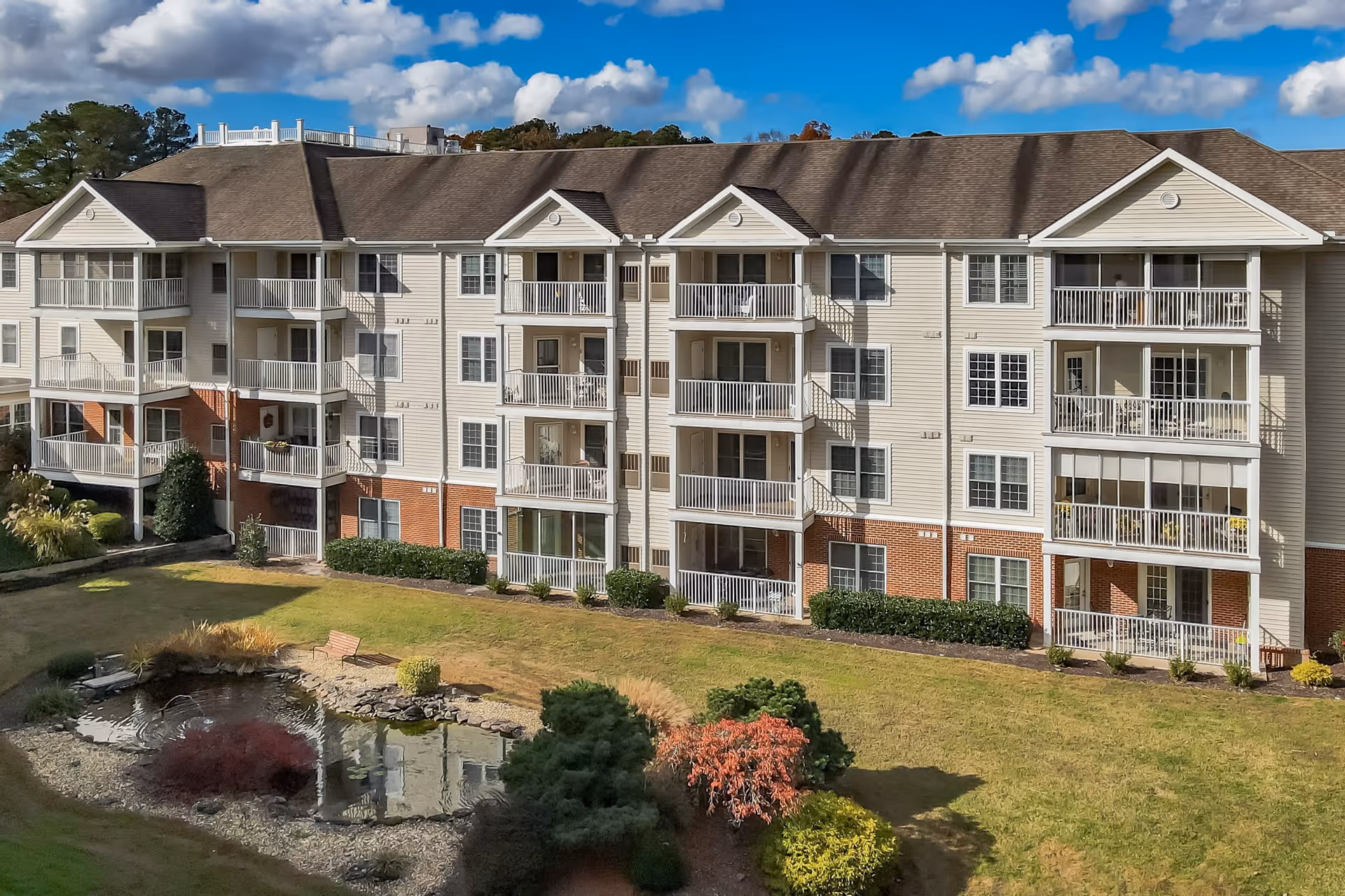 Exterior view of a multi-story senior living facility building with balconies, surrounded by a landscaped garden featuring a small pond, shrubs, and a bench under a partly cloudy blue sky.
