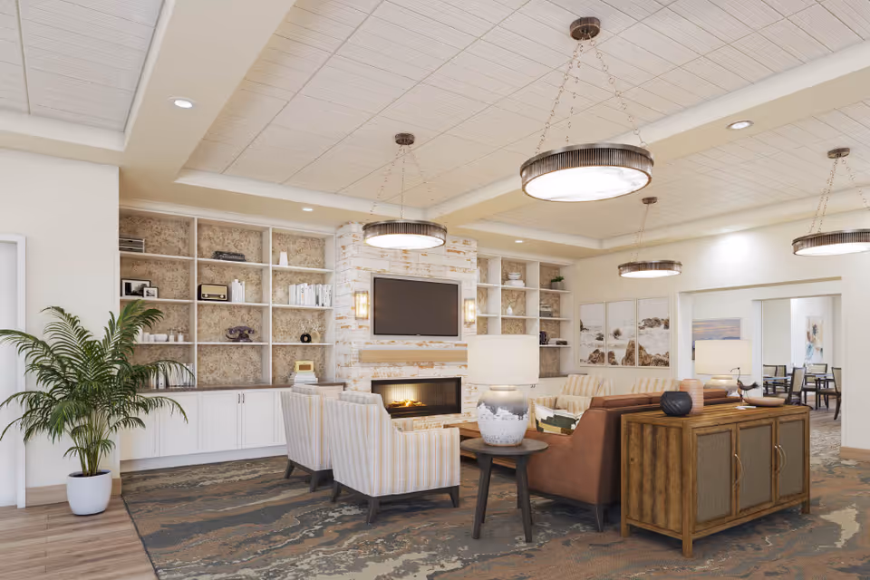 A spacious and well-lit living room area in a senior living facility featuring a modern fireplace with a mounted flat-screen TV above it. The room has built-in shelves filled with decorative items and books, comfortable seating including striped armchairs and a brown leather sofa, a wooden sideboard with a lamp and decorative objects, and large circular ceiling lights. There is a large potted plant on the left side and artwork on the walls in the background.