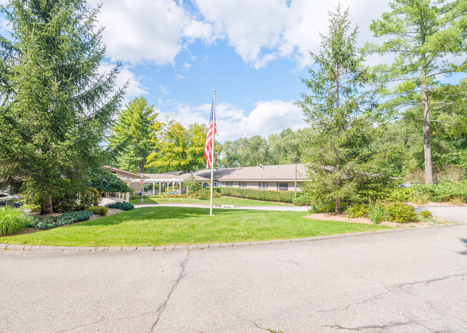 Exterior view of The Country House in Westchester facility showing a single-story building surrounded by green trees and shrubs under a partly cloudy blue sky. An American flag is prominently displayed on a flagpole in the center of a grassy area in front of the building. A paved driveway curves around the grassy area.