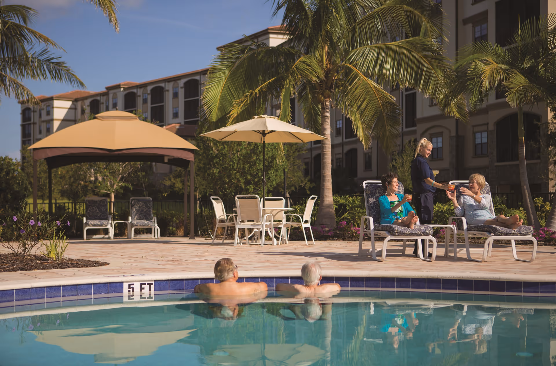 Two elderly men relaxing in a swimming pool while two elderly women sit on lounge chairs by the poolside, being served drinks by a caregiver. The scene is set outdoors with palm trees, patio umbrellas, and a multi-story residential building in the background under a clear blue sky.