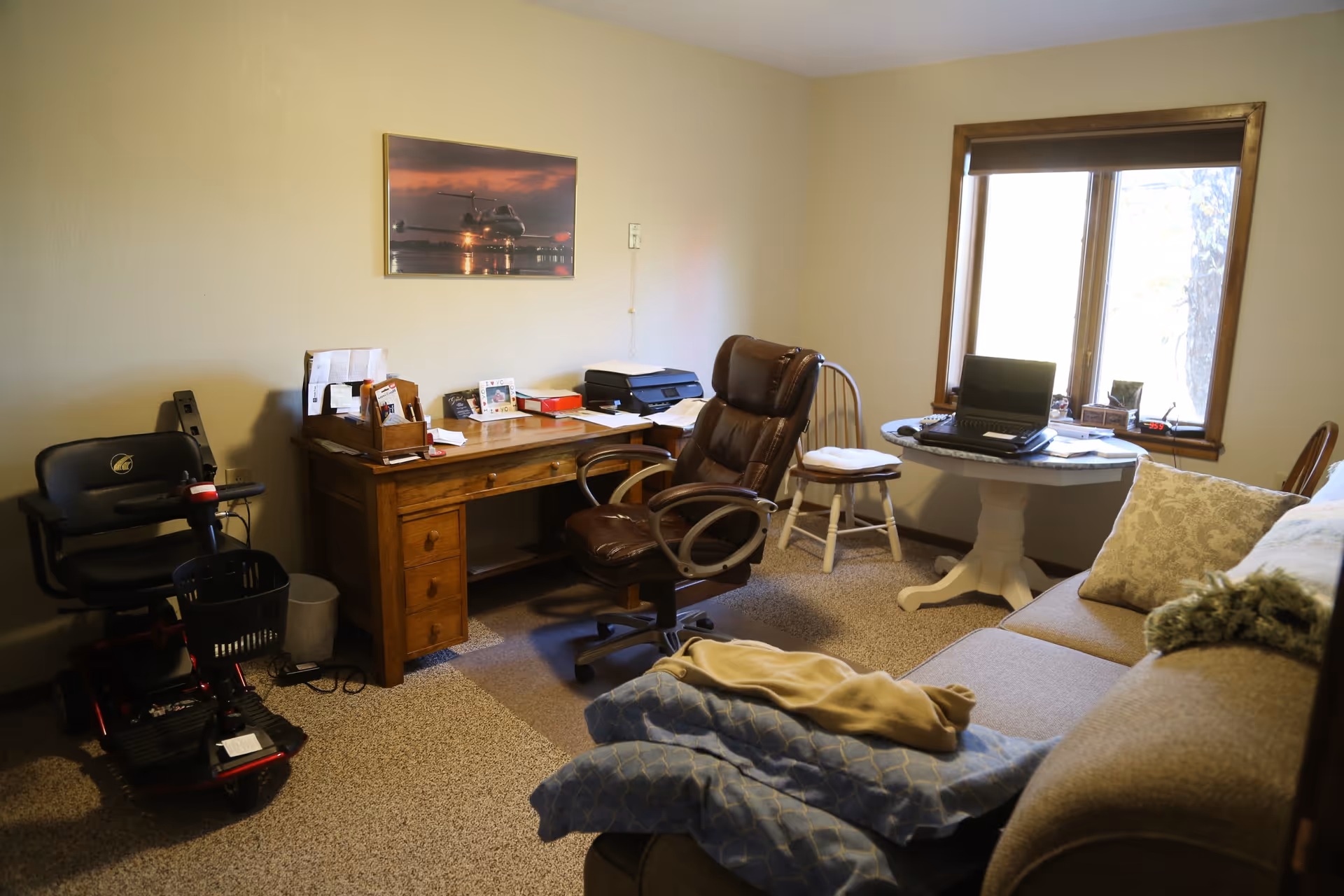 A cozy room with a wooden desk holding papers, a printer, and office supplies. A brown leather office chair is in front of the desk. To the right, there is a small round table with a laptop and some books, positioned near a window with a brown blind. A cushioned wooden chair is next to the table. In the foreground, a beige sofa with patterned pillows and a folded blanket is visible. A mobility scooter is parked to the left of the desk. The walls are light-colored, and a framed picture of an airplane at sunset hangs above the desk.