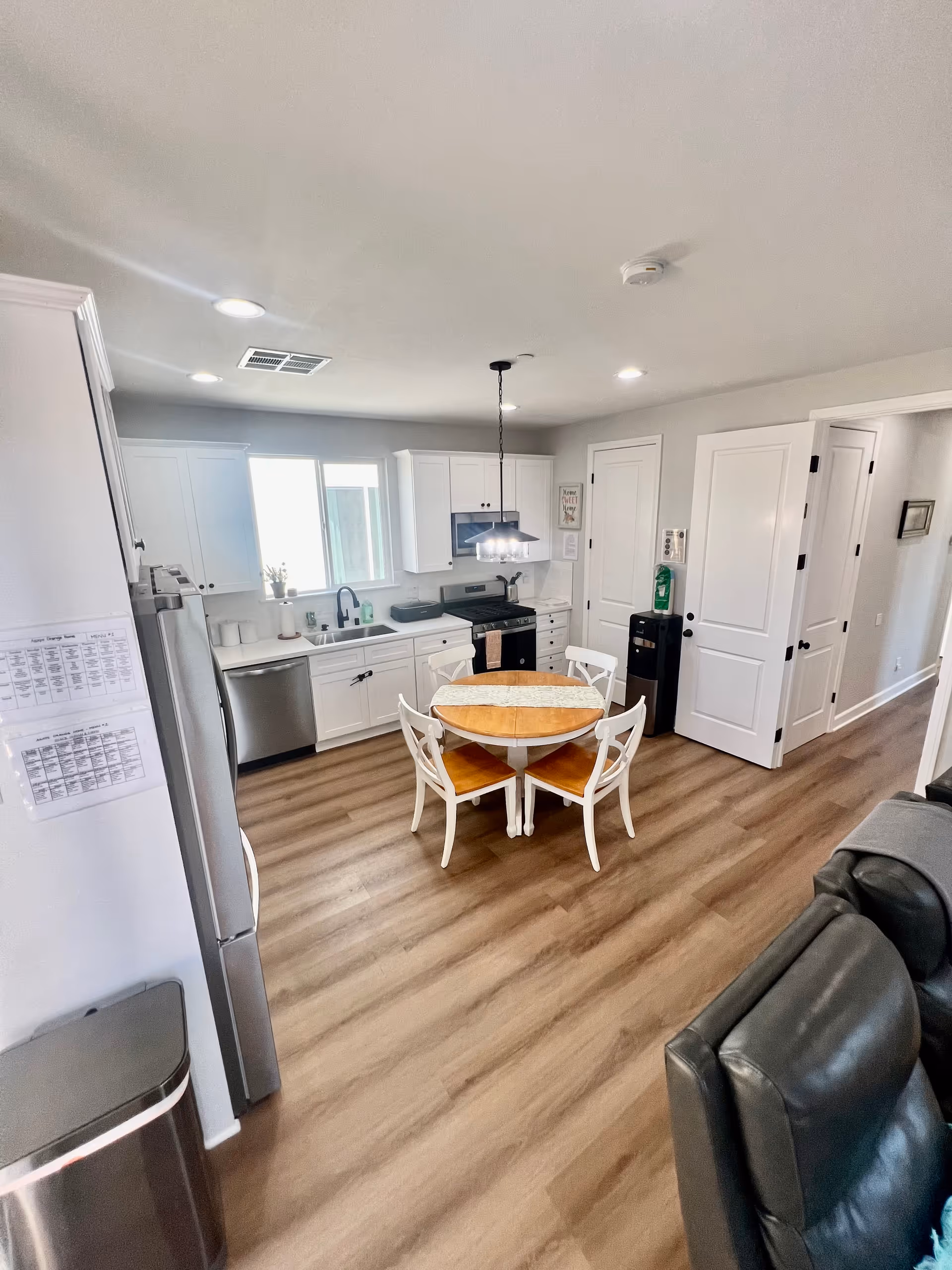 A bright kitchen and dining area with white cabinets, stainless steel appliances including a dishwasher and stove, a round wooden dining table with four white chairs, wood flooring, and a ceiling light fixture. There is a window above the sink and two closed white doors on the right side. Part of a black leather couch is visible in the foreground.