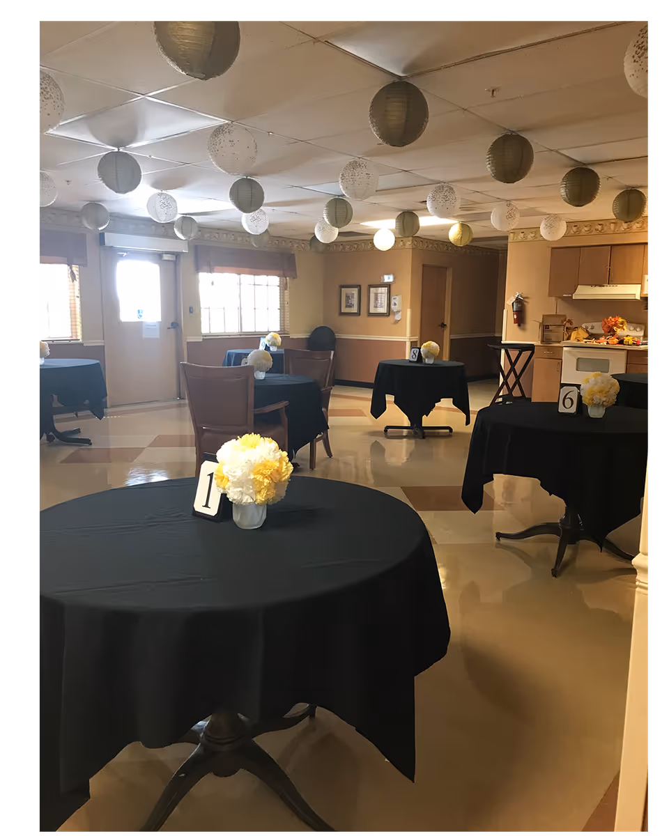 A decorated dining room with round tables covered in black tablecloths, numbered floral centerpieces, chairs, and hanging paper lanterns.