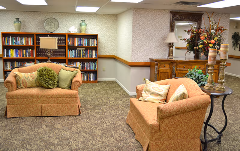A cozy sitting area in a senior living facility with two upholstered armchairs adorned with decorative pillows. Behind the chairs is a wooden bookshelf filled with books and decorative items, including vases and a lamp. To the right, there is a wooden sideboard with a large mirror above it and a floral arrangement, along with two tall candles on a small round table. The room has patterned carpet and wallpaper with soft lighting from ceiling fixtures.