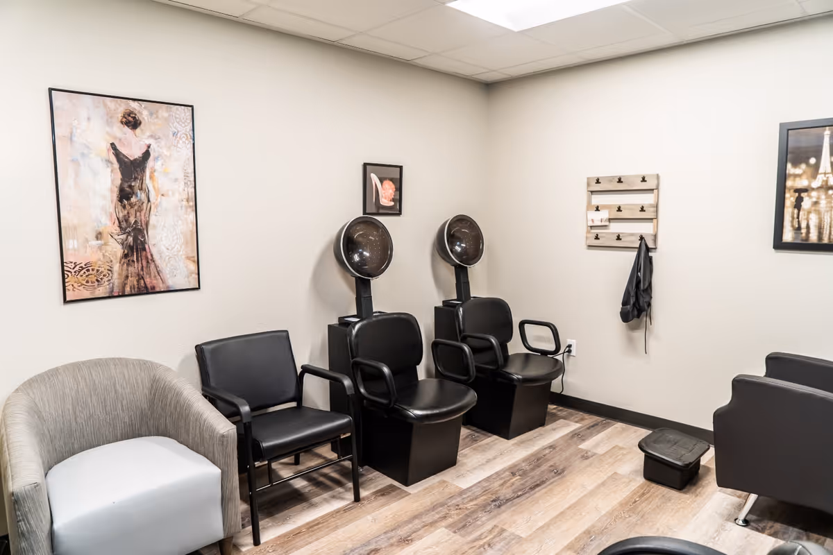 Interior room with two black hair drying chairs, two additional chairs, a small black stool, and wall decorations including framed pictures and a wooden wall organizer with hooks.