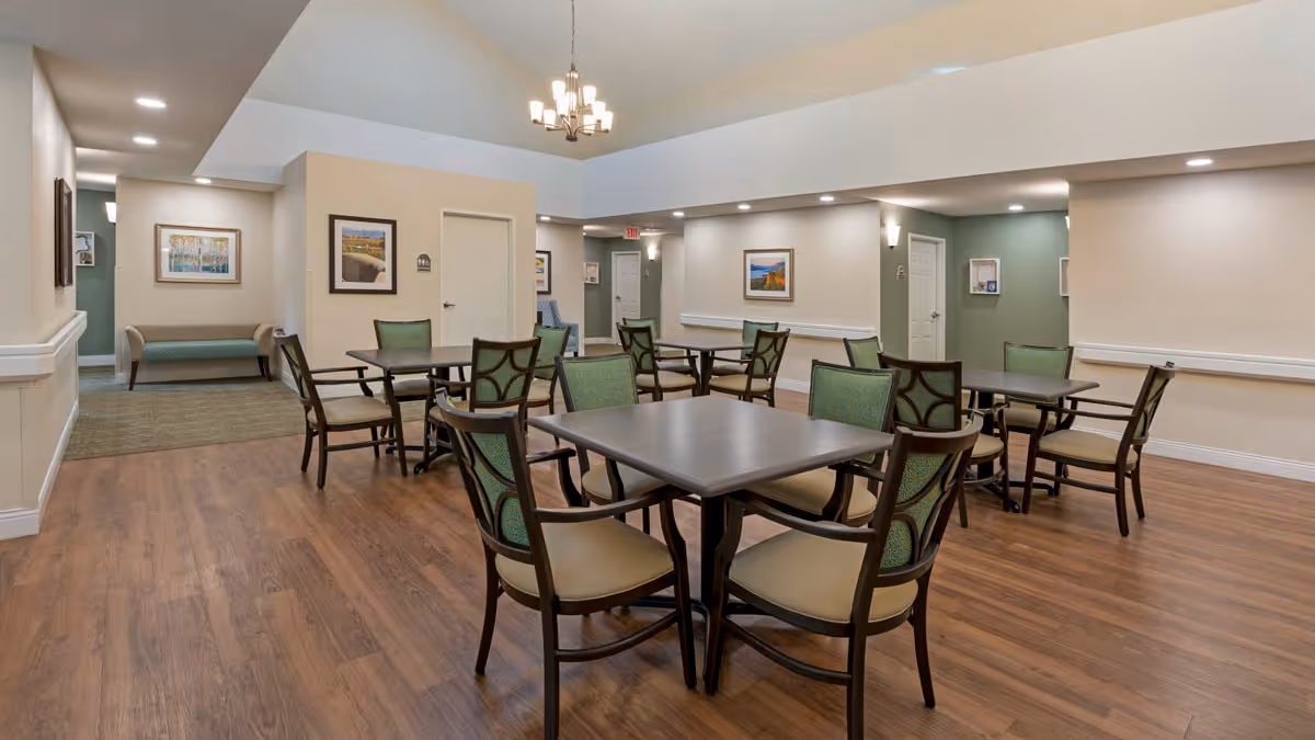 A spacious dining area with several square tables and green cushioned chairs arranged neatly on a wooden floor. The room has beige walls with framed artwork, recessed lighting, and a chandelier hanging from the ceiling. There is a hallway with a bench and additional seating visible in the background.
