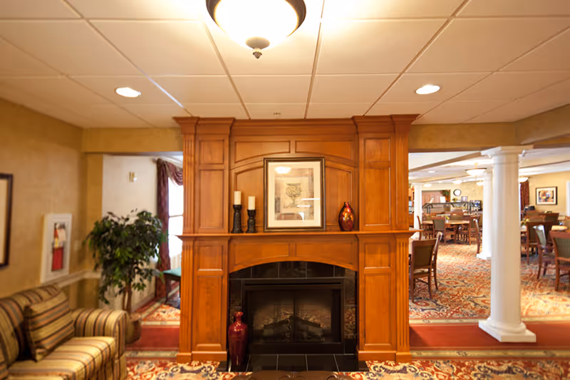A cozy interior space featuring a wooden fireplace mantel with decorative items including candles, a framed picture, and vases. To the left, there is a striped armchair and a potted plant. In the background, a dining area with tables and chairs is visible, along with white columns and warm lighting.