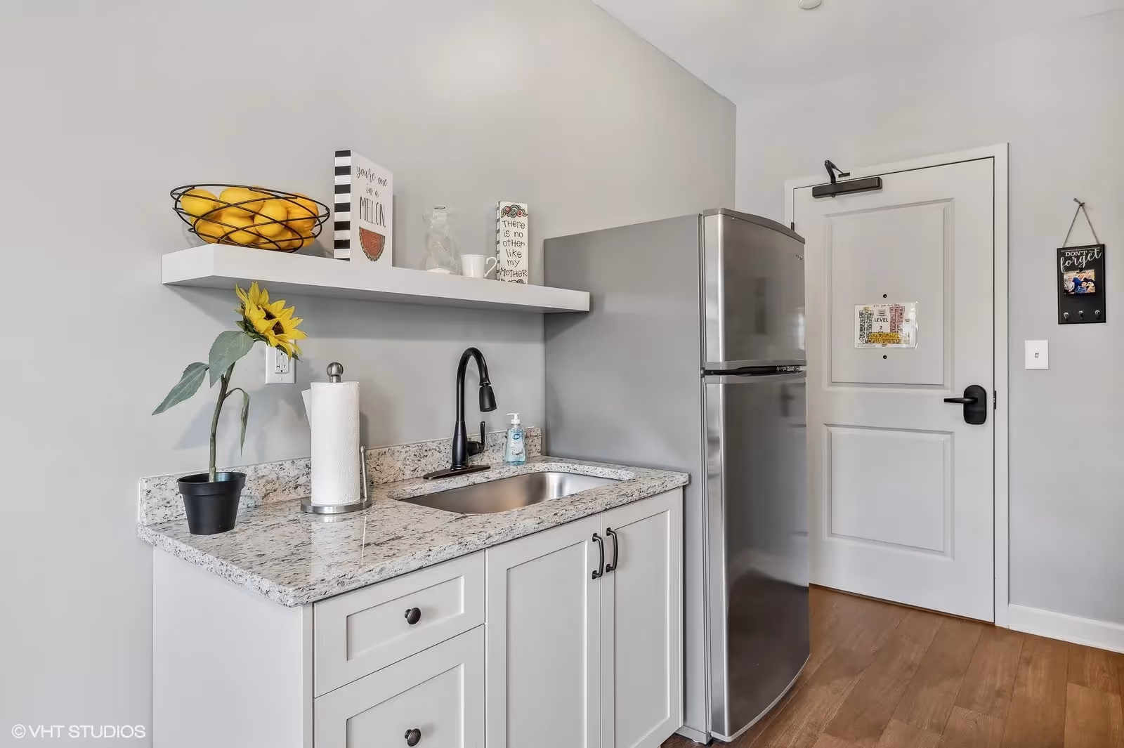 Compact kitchenette featuring a granite countertop with sink, floating shelf with decor, stainless-steel refrigerator, and an entry door.