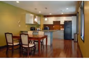 Open-plan dining area with a wooden table and chairs facing a modern kitchen with an island, pendant lights, and hardwood floors.