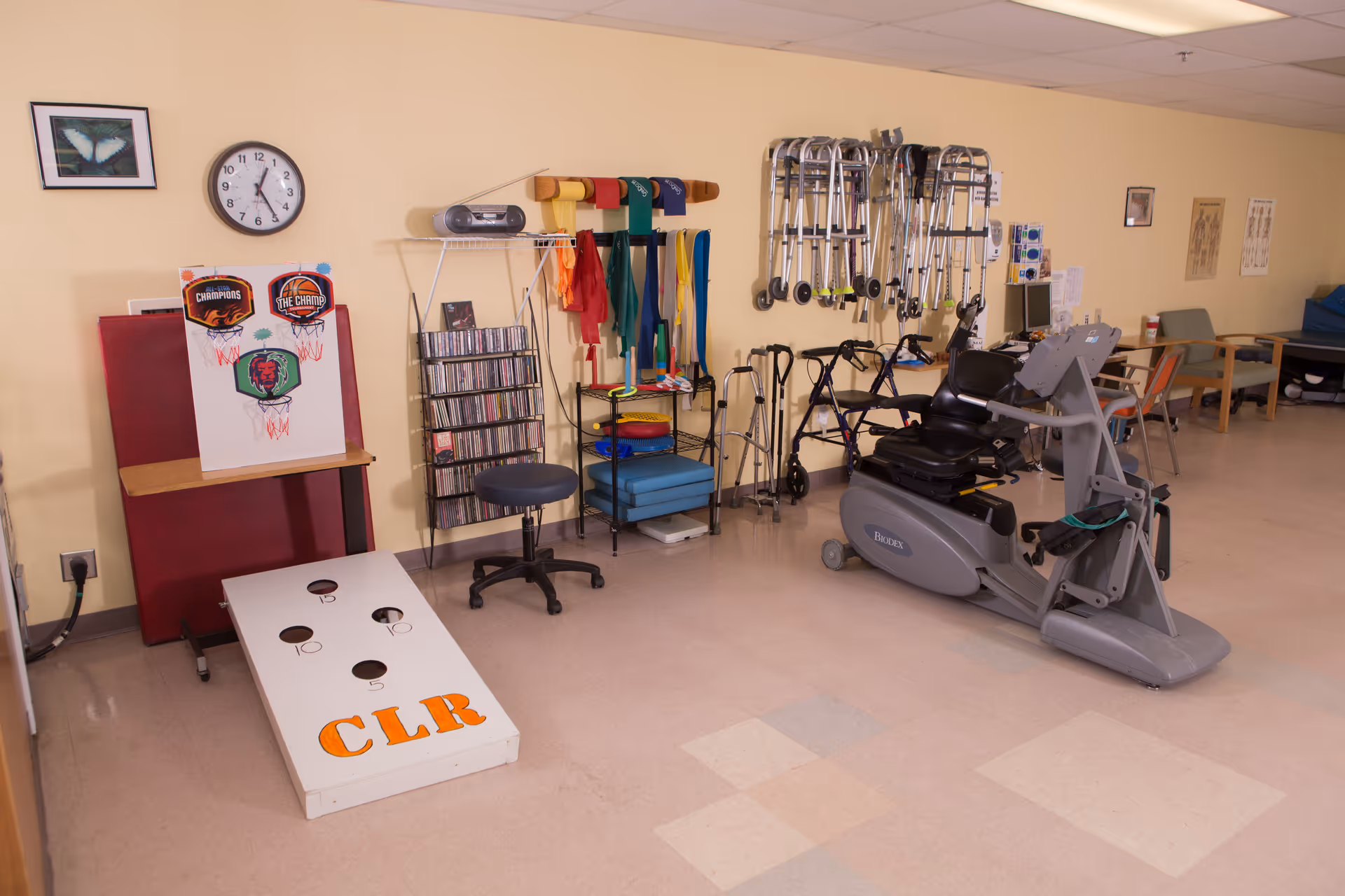 Rehabilitation therapy room containing an exercise bike, walkers and mobility aids, shelves of therapy equipment, and a cornhole board labeled CLR.