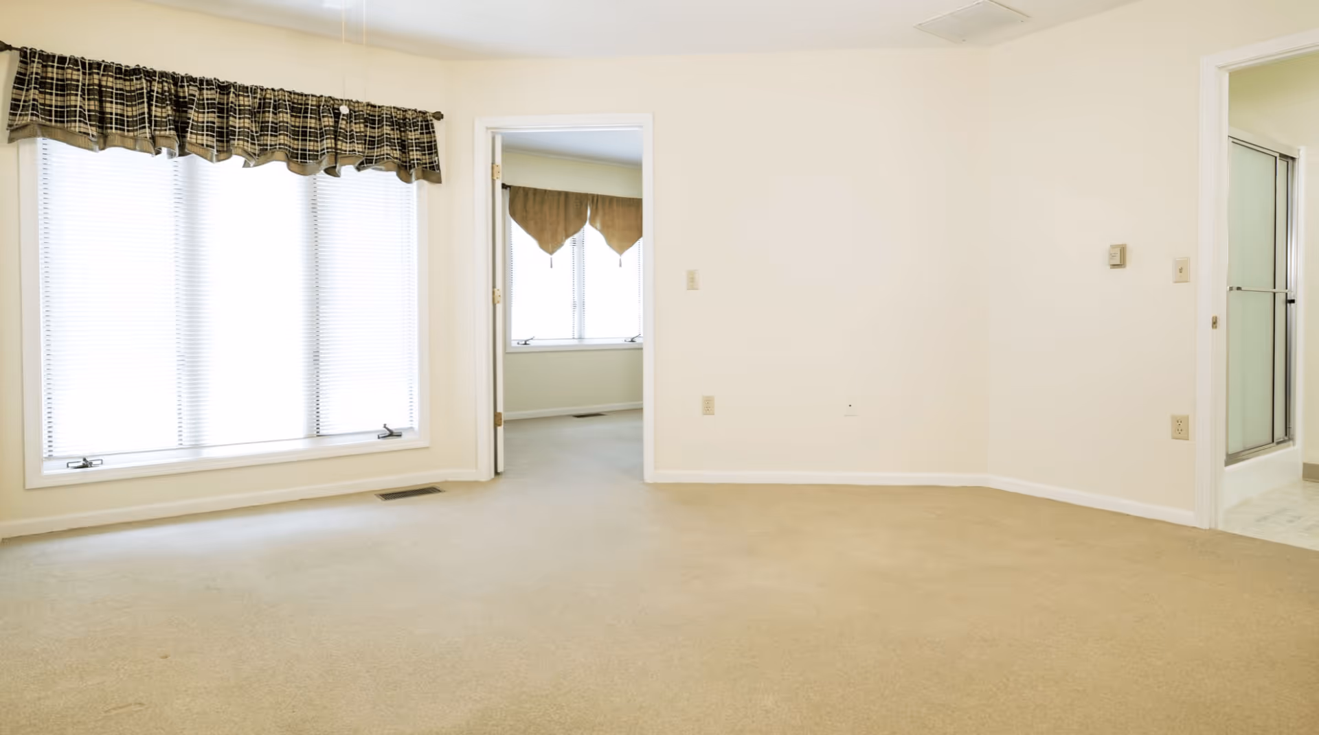 Empty room with beige carpet and cream-colored walls. A large window with blinds and a plaid valance is on the left wall. There are two doorways: one in the center leading to another room with a window and valance, and one on the right leading to a bathroom with a glass shower door.