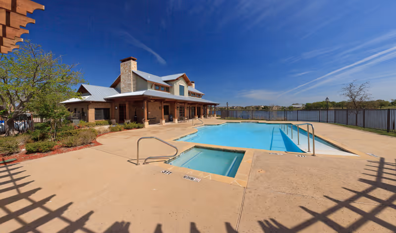Outdoor view of a swimming pool and hot tub beside a lakeside clubhouse under a clear blue sky.