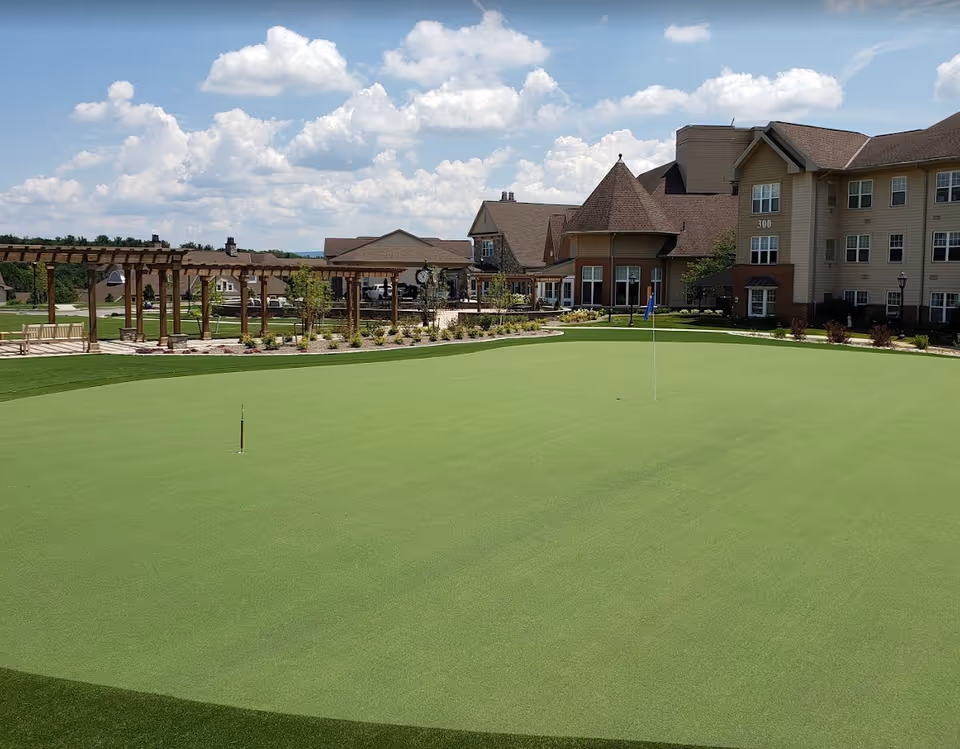 Outdoor putting green with two golf holes and flags in front of a senior living facility building under a partly cloudy sky. The building has a beige exterior with multiple windows and a wooden pergola with benches and landscaping around the green.