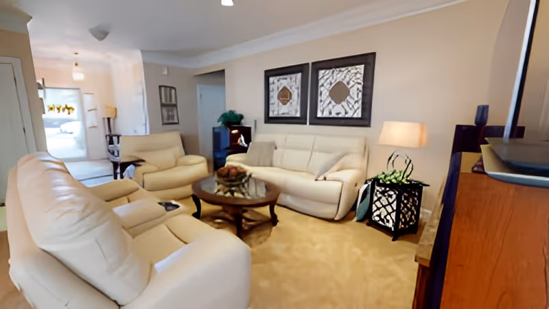 A cozy living room with beige leather sofas and armchairs arranged around a round wooden coffee table. The walls are light-colored with two framed artworks hanging above the sofa. A small side table with a lamp and a decorative plant is placed next to the sofa. The room has a carpeted floor and a TV on a wooden stand is visible on the right side.