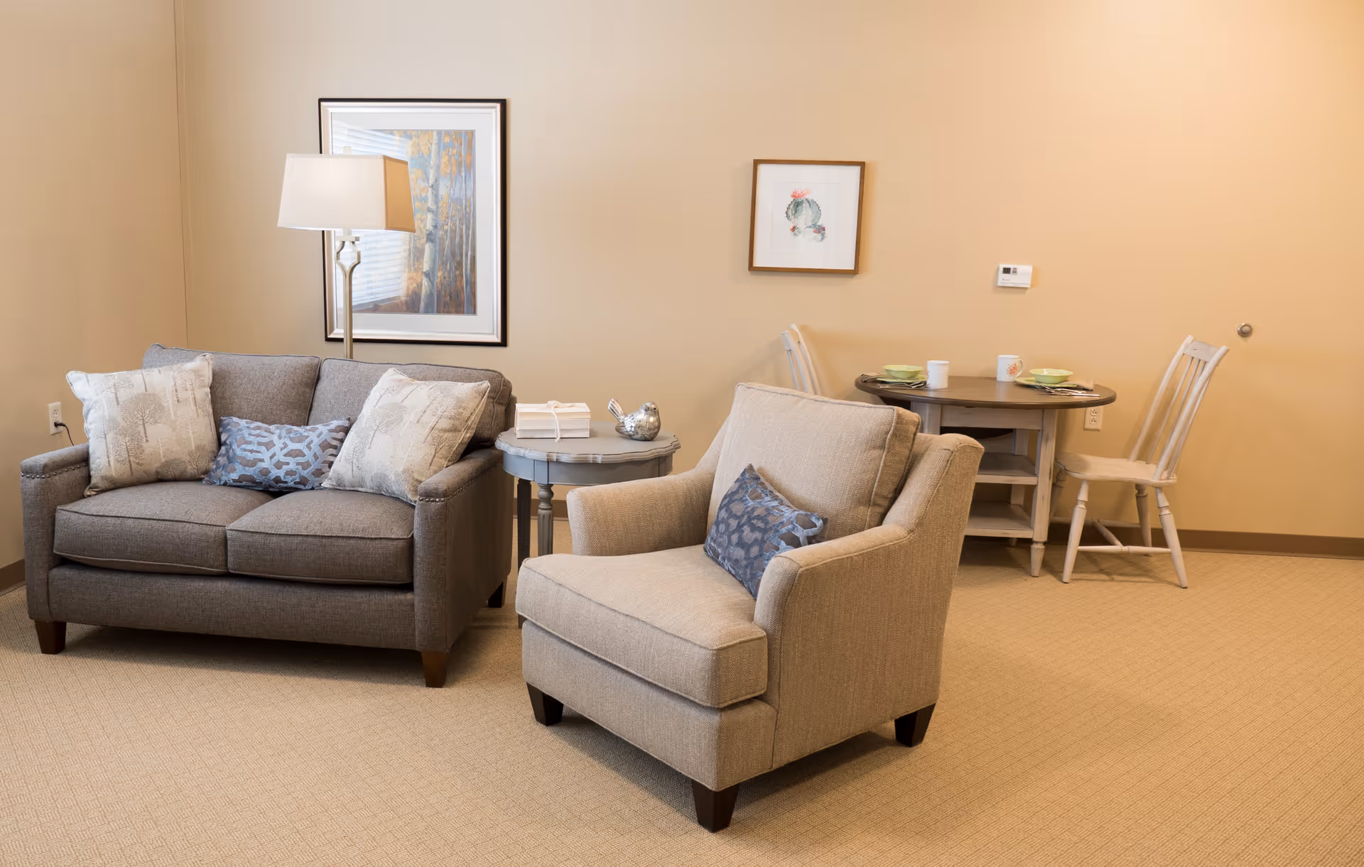 Neutral-toned living area with a gray loveseat, beige armchair, side table with lamp, and a small dining table against a beige wall.