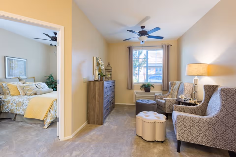 A cozy living area with two patterned armchairs, a small round table with a lamp, and two ottomans. A window with beige curtains lets in natural light. To the left, an open doorway reveals a bedroom with a bed covered in yellow and white floral bedding and a ceiling fan.