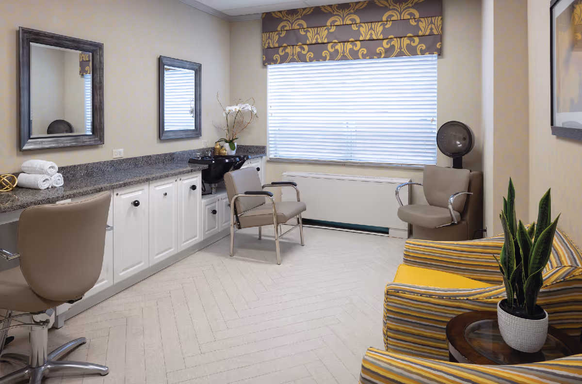 A bright and clean salon room with two mirrors above a granite countertop with white cabinets underneath. There are three chairs: one salon chair, one gray chair in front of a black hair washing sink, and another gray chair with a hair dryer hood. A large window with white blinds and a decorative valance lets in natural light. A striped yellow and gray armchair with a small round table and a potted plant is in the foreground.