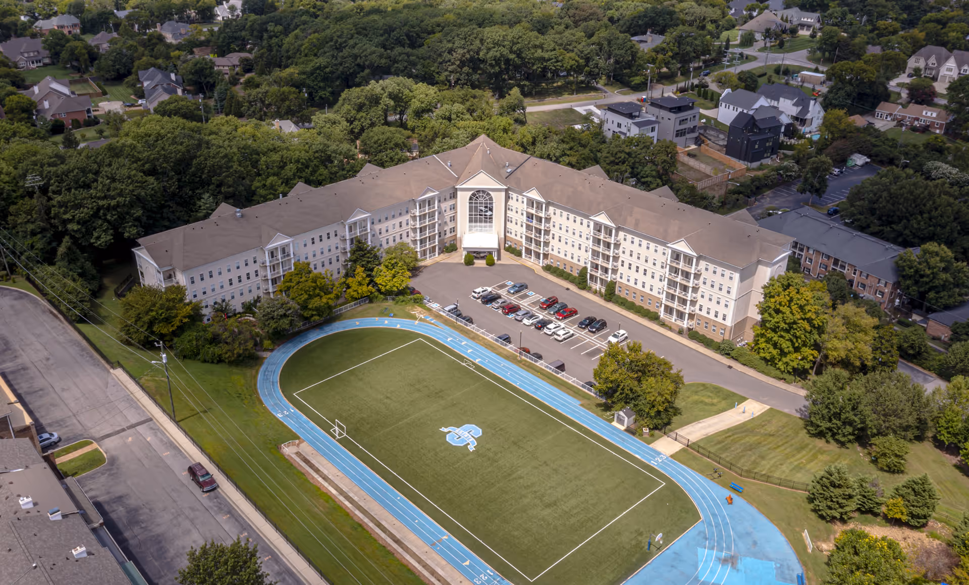 Aerial view of The St. Paul Senior Living Community building surrounded by trees and residential houses, with a parking lot in front and a sports field with a blue running track adjacent to the building.