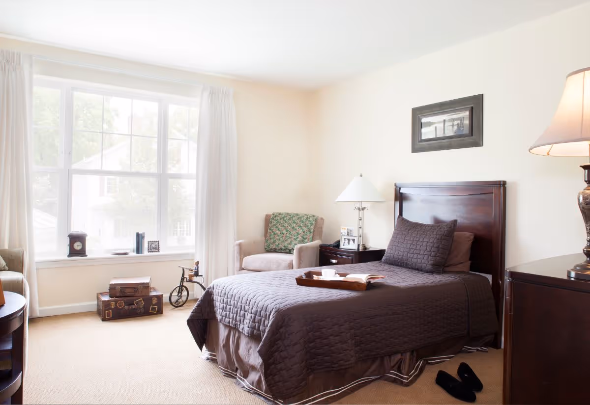 A bright and cozy bedroom with a single bed covered in a dark quilted bedspread. There is a wooden headboard, a bedside table with a lamp and framed photos, a comfortable armchair with a patterned cushion, and a large window with white curtains letting in natural light. A tray with a cup and an open book rests on the bed, and a pair of black slippers is on the carpeted floor.