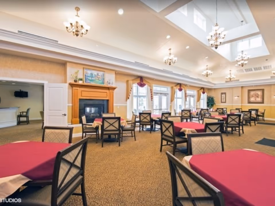Large bright dining room with multiple tables covered in red tablecloths, chandeliers, and a fireplace.