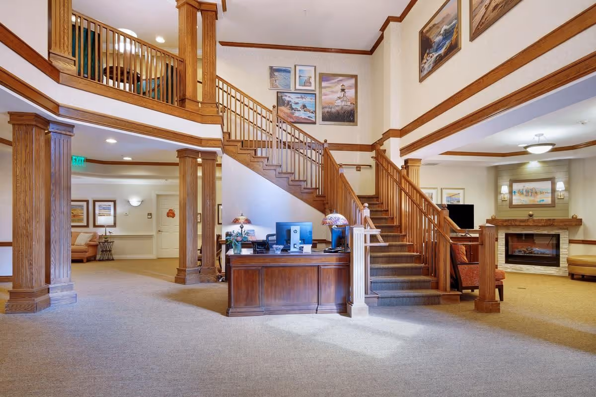 Spacious senior living lobby with a wooden reception desk, central staircase, seating areas and a fireplace.