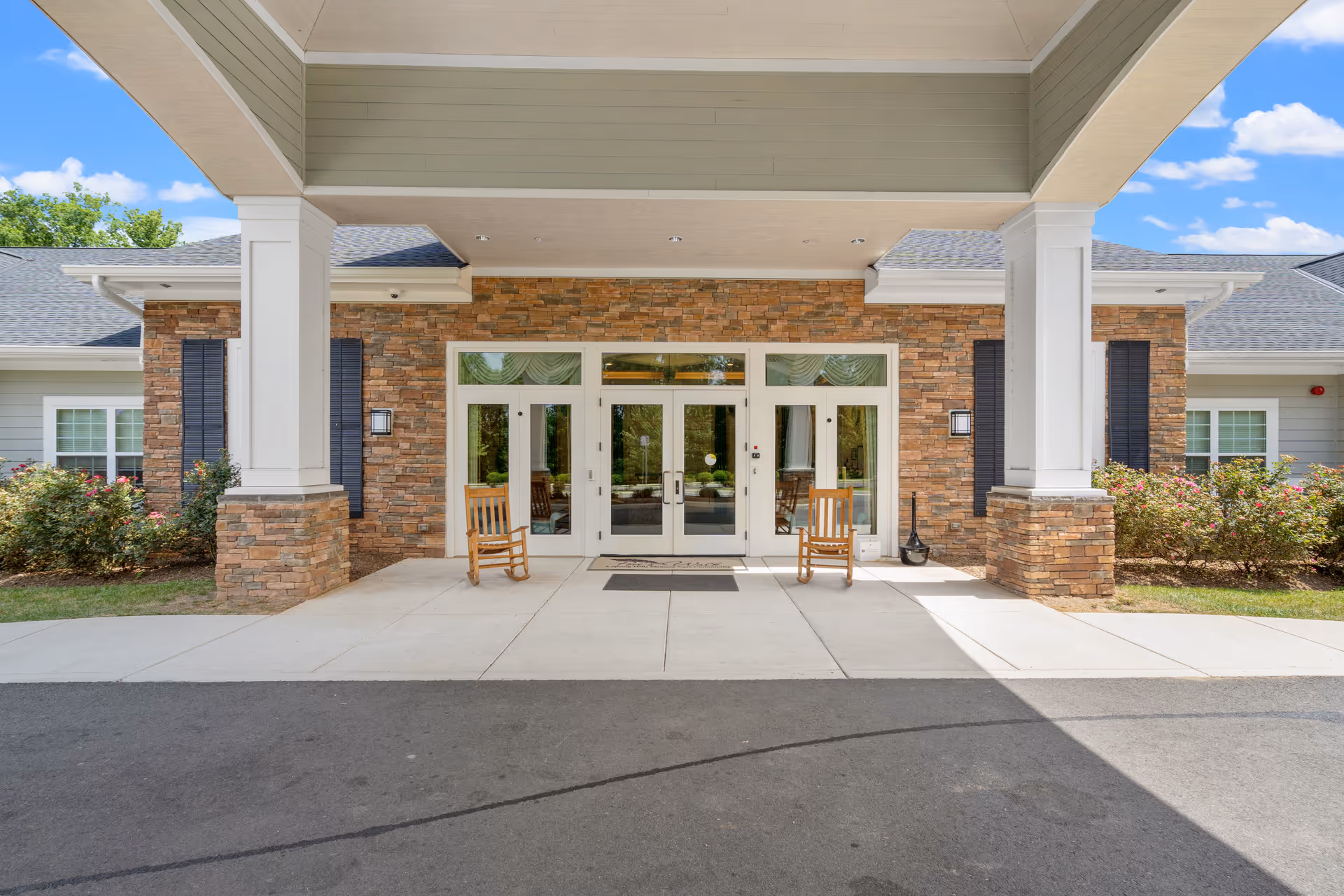 Entrance of a senior living facility with a covered driveway, two wooden rocking chairs on either side of double glass doors, brick and siding exterior walls, and landscaping with bushes and flowers under a blue sky with some clouds.