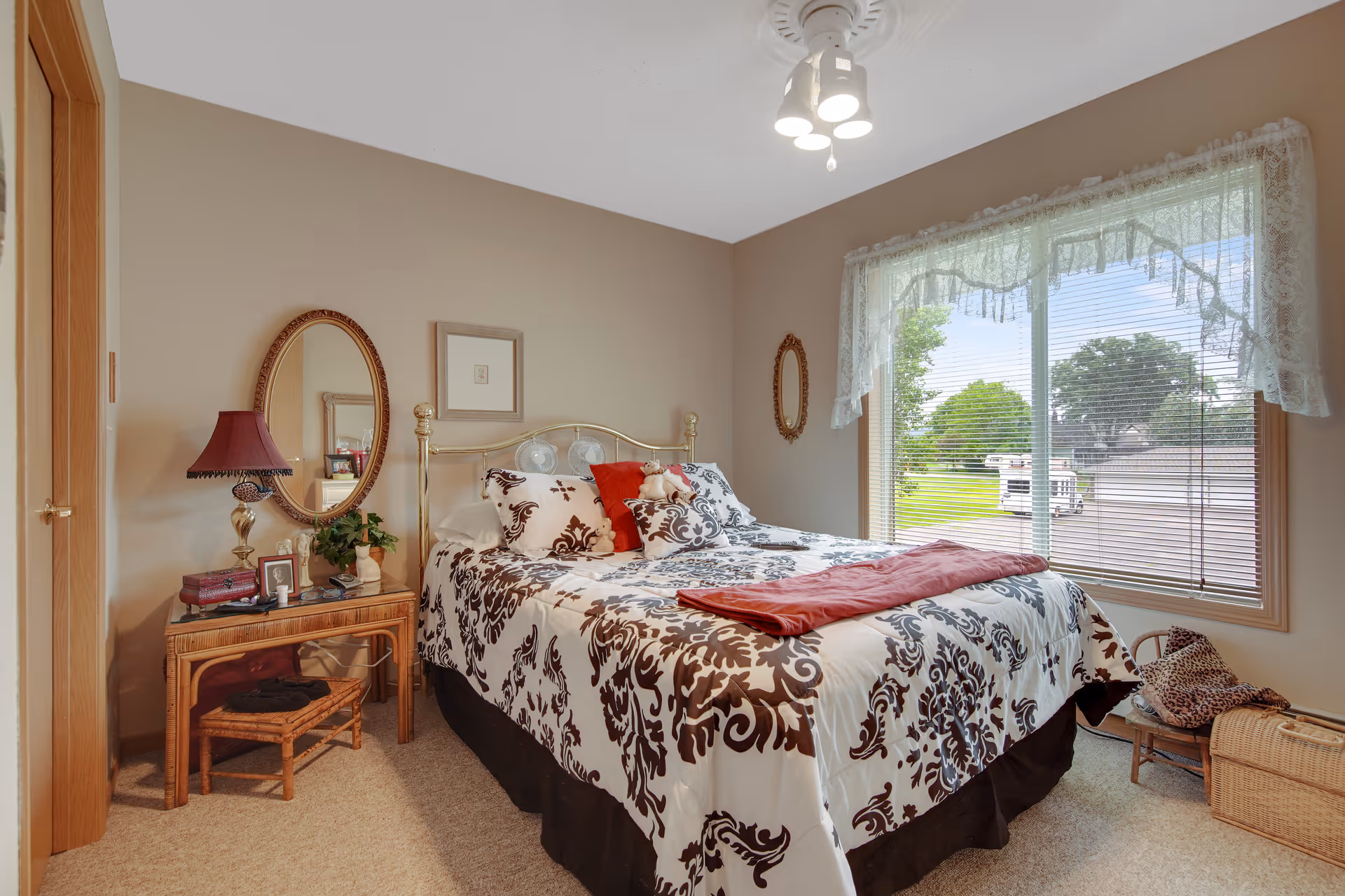 Sunlit bedroom with a decorated double bed with patterned bedding, a nightstand with lamp and mirror, and a large window overlooking a green lawn.