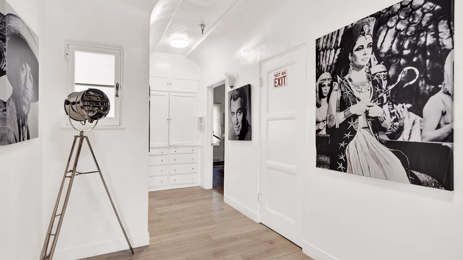 A bright hallway with light wooden flooring and white walls. The hallway features black and white portraits on the walls, a vintage-style tripod floor lamp, a small window, and a door with a 'NOT AN EXIT' sign. Built-in white cabinets are visible at the end of the hallway.